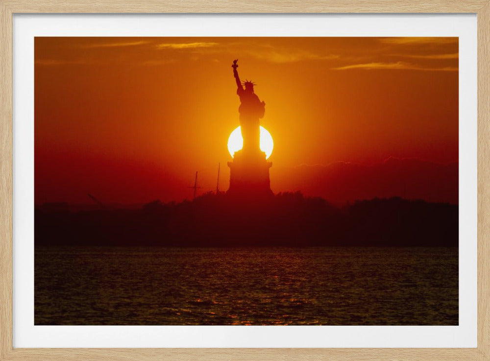 A powerful silhouette of the Statue of Liberty against a fiery orange and red sunset. The sun is perfectly positioned behind the statue's base, creating a brilliant, glowing orb of light. The dark waters in the foreground reflect the vibrant colors of the sky. The photograph is presented within a silver-colored frame. Artwork