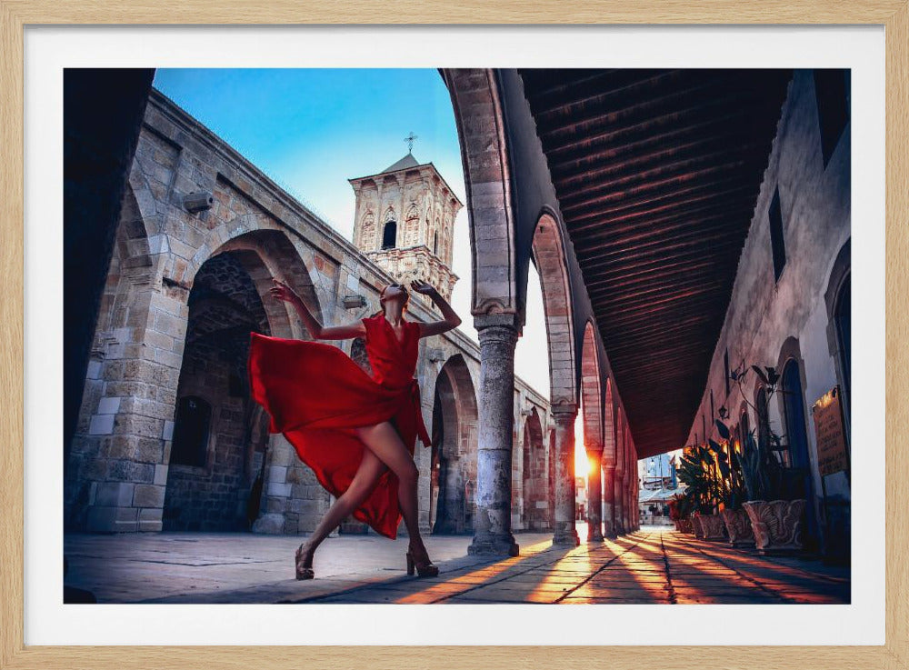 A woman in a vibrant, flowing red dress strikes a dramatic pose in a stone colonnade at sunset. The low sun casts long, golden shadows on the ground. In the background, a stone church tower stands against a clear blue sky. Artwork