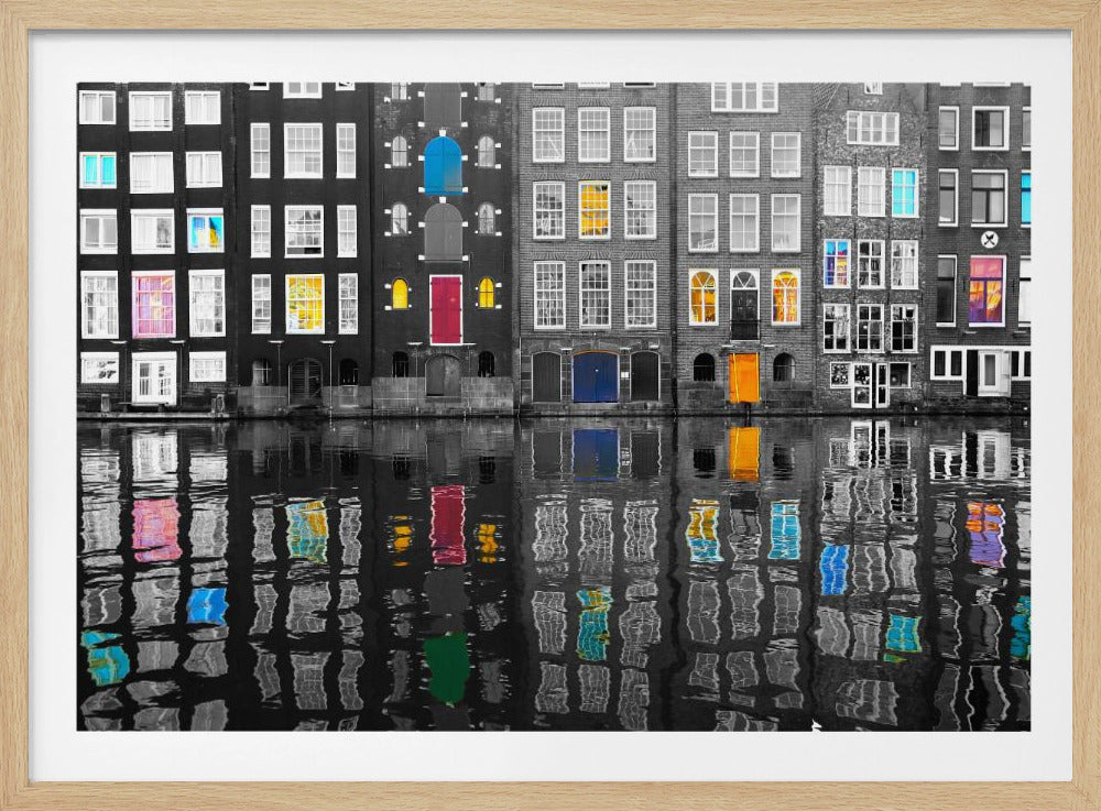 A framed black and white photograph of canal houses in Amsterdam, with selective coloring highlighting windows and doors in vibrant shades of red, blue, yellow, and pink. The colorful architectural details are mirrored in the rippling water of the canal below. Artwork