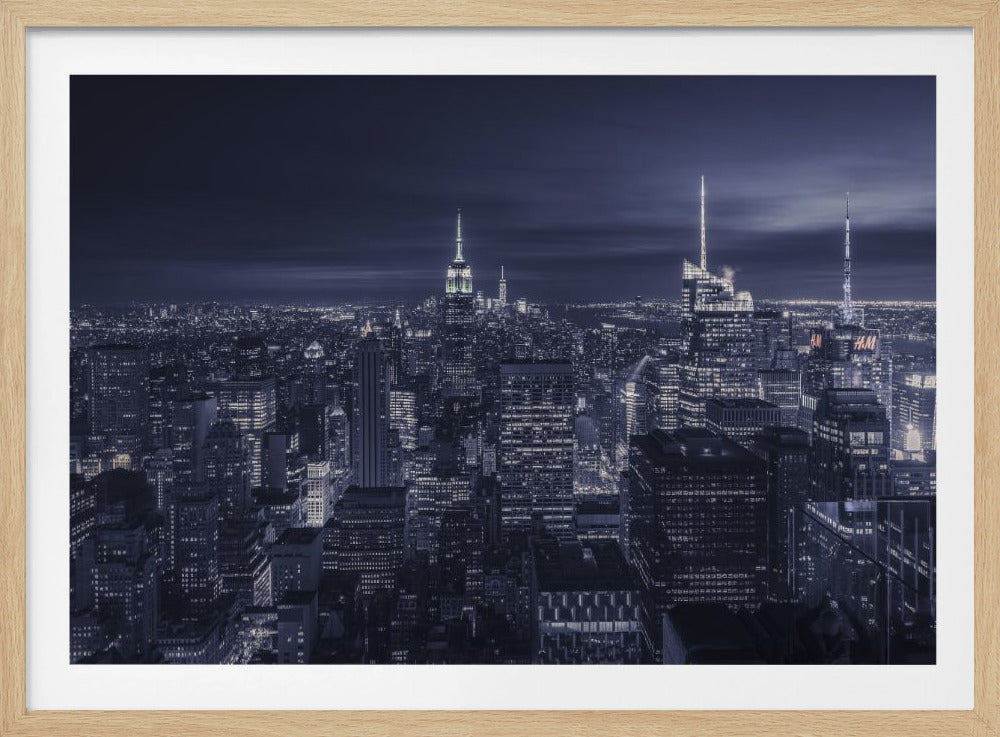 A dramatic nighttime photograph of the New York City skyline, viewed from a high vantage point. The cityscape is a sea of illuminated skyscrapers under a dark, moody sky, with the entire image cast in a cool blue and white monochrome tone and presented in a silver frame. Artwork
