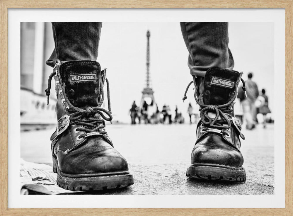 A low-angle, black and white photograph focusing on a pair of well-worn Harley-Davidson combat boots on a city street. In the blurred background, the Eiffel Tower and the silhouettes of people can be seen, creating a sense of travel and urban exploration. Decor