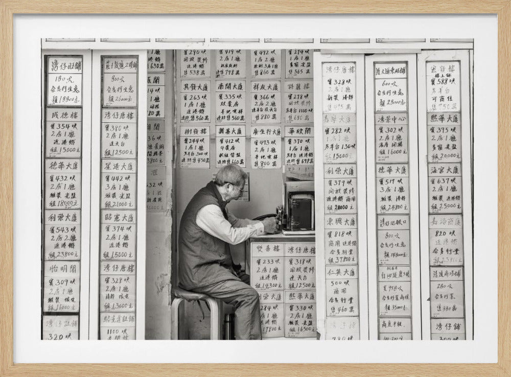 A black and white photograph of an elderly man with glasses sitting on a stool in a small, cramped shop. He is hunched over a counter, operating a small machine. The walls around him are completely covered with vertical paper signs featuring handwritten Chinese calligraphy, likely property listings or advertisements. Artwork