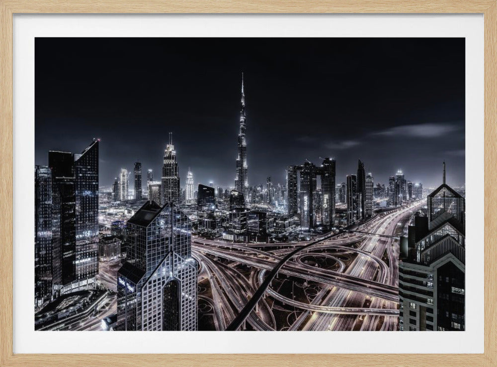 A striking black and white aerial photograph of the Dubai skyline at night, captured in a silver frame. The Burj Khalifa stands tall in the background, while a complex web of illuminated highway overpasses and light trails from traffic fills the foreground, creating a dynamic and futuristic cityscape. Print