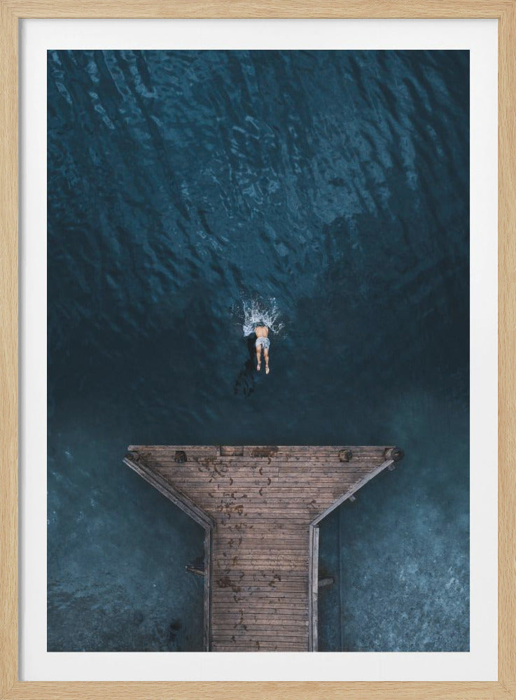 An aerial top-down photograph of a lone person swimming in deep, dark blue water, just off the edge of a weathered wooden pier. The swimmer creates a small splash as they move away from the dock. Wall Art