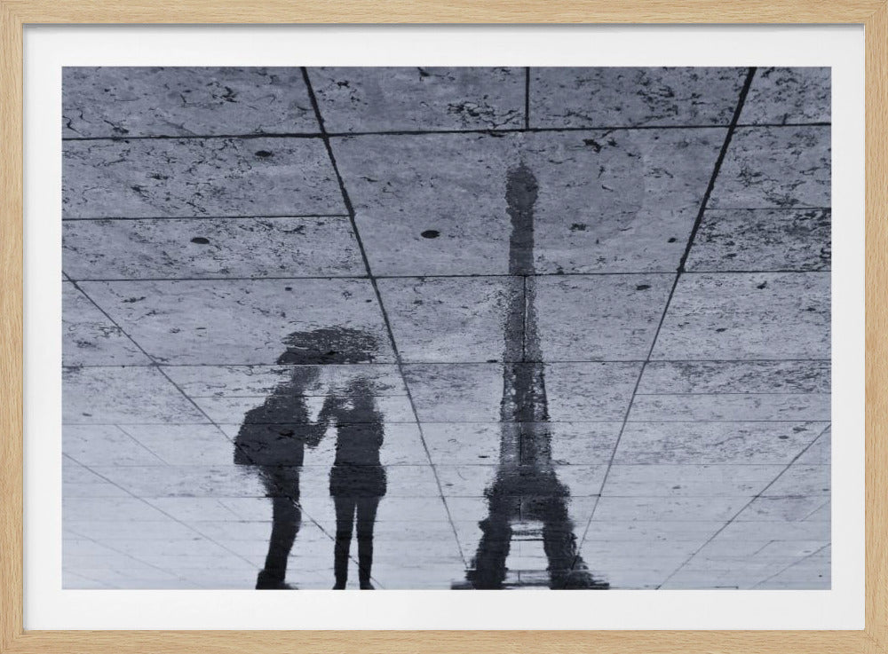 A framed, black and white photograph capturing the reflection of the Eiffel Tower and a romantic couple under an umbrella on wet, tiled pavement. Print