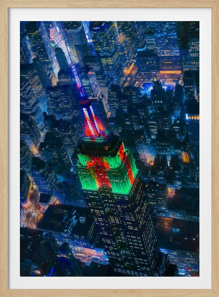 A dramatic high-angle aerial view of the Empire State Building at night, illuminated in festive red and green lights. The surrounding New York City skyline is a dense grid of skyscrapers glowing with blue and white lights, creating a vibrant, sprawling cityscape. Poster