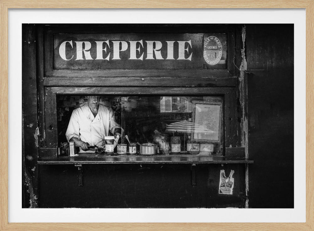 A classic black and white photograph of a Parisian crêperie stand, framed in silver. An elderly man in a chef's uniform is seen through the service window preparing a crêpe, with a prominent 'CREPERIE' sign above him. Decor
