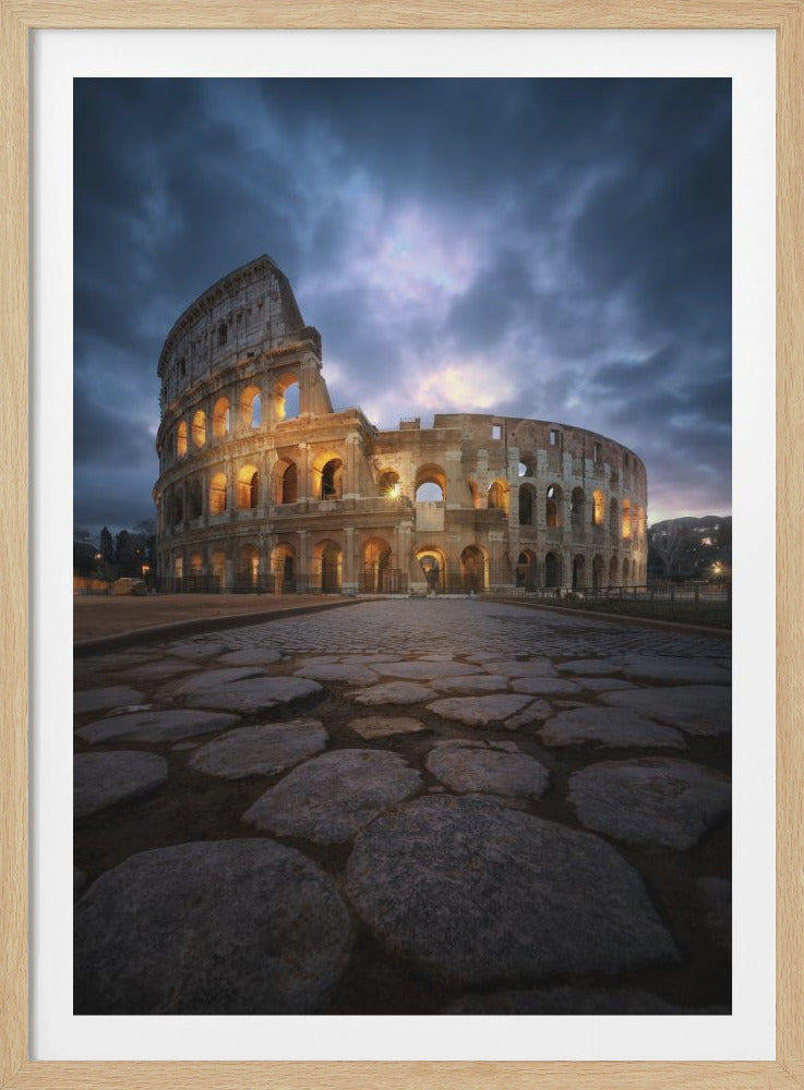 A dramatic low-angle photograph of the Colosseum in Rome at dusk, with its ancient arches illuminated by warm golden lights under a stormy, dark blue sky. A weathered cobblestone road fills the foreground, leading the viewer's eye to the historic landmark. Poster