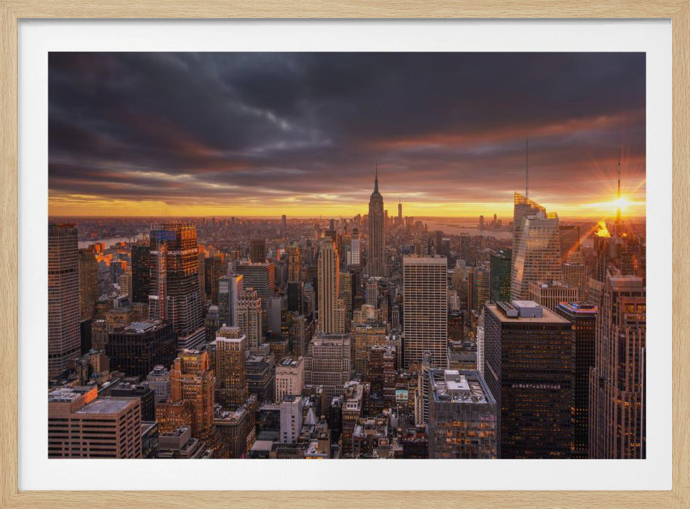 A dramatic framed photograph of the New York City skyline at sunset. The iconic Empire State Building is visible in the center as a brilliant orange sunburst on the horizon contrasts with dark, stormy clouds overhead, illuminating the city. Print