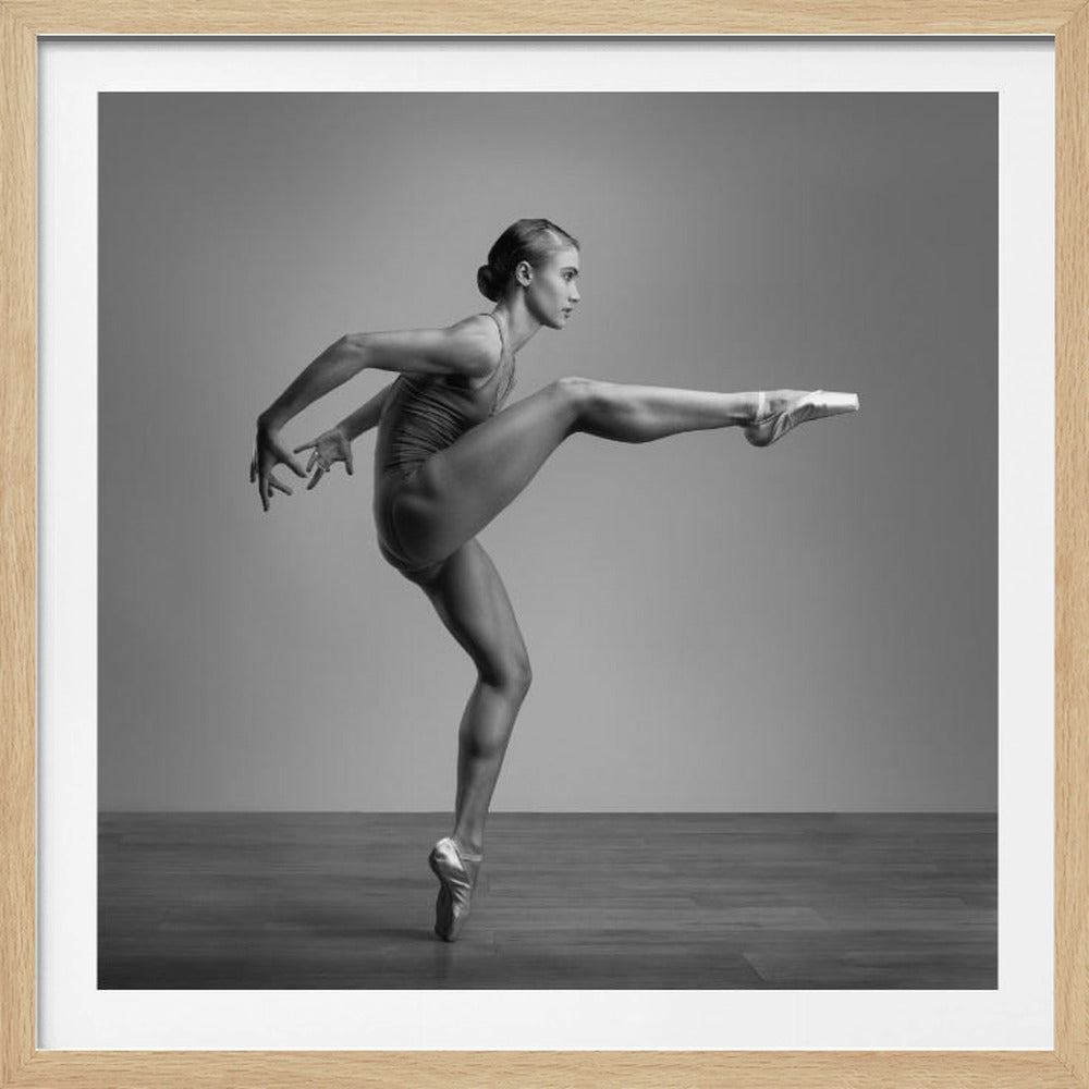 A black and white studio photograph of a muscular female ballet dancer in a dynamic pose, set within a light wood frame. She balances on one pointe shoe, with her other leg extended straight out to the side at a high angle. Her body is turned in profile, showing her strength and grace against a plain grey background and dark wood floor. Decor