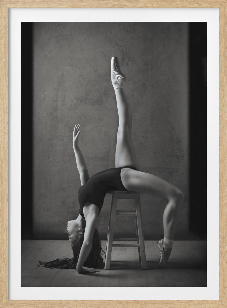 A black and white studio photograph of a graceful ballerina in a black leotard and pointe shoes, executing a dramatic backbend over a wooden stool. One leg extends vertically into the air while her head and long hair hang towards the floor, showcasing her incredible flexibility and strength against a simple, textured gray background. Decor