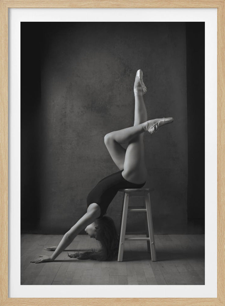 A black and white studio photograph of a graceful ballerina in a dramatic pose. She wears a leotard and pointe shoes, bending over a wooden stool with her legs extended vertically in the air, showcasing her flexibility and strength against a dark, moody background. Print
