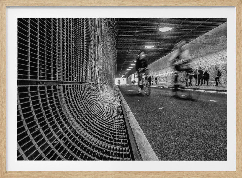 A dynamic black and white photograph from inside a modern tunnel. On the left, a curved metal grate wall creates a strong pattern, while on the right, cyclists are captured in motion blur on a paved path. Pedestrians are visible in the background, walking through the illuminated passage. Poster