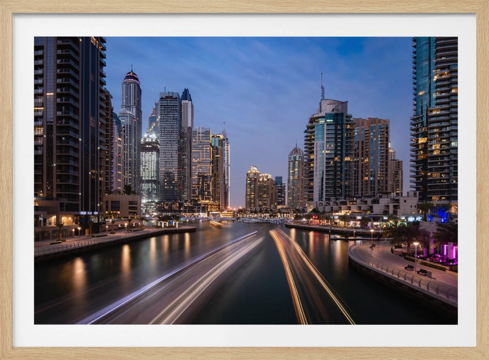 A long-exposure photograph of the Dubai Marina at dusk, framed in silver. Illuminated skyscrapers line a busy waterway, with light trails from moving boats creating long, bright streaks on the water. The sky is a deep twilight blue, and the city's lights cast a warm glow. Decor
