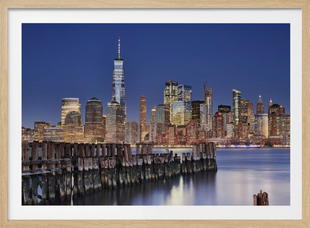 A framed photograph of the Lower Manhattan skyline at dusk. The One World Trade Center and other skyscrapers are brightly lit against a deep blue sky. In the foreground, an old wooden pier extends into the water, which reflects the city lights. Wall Art
