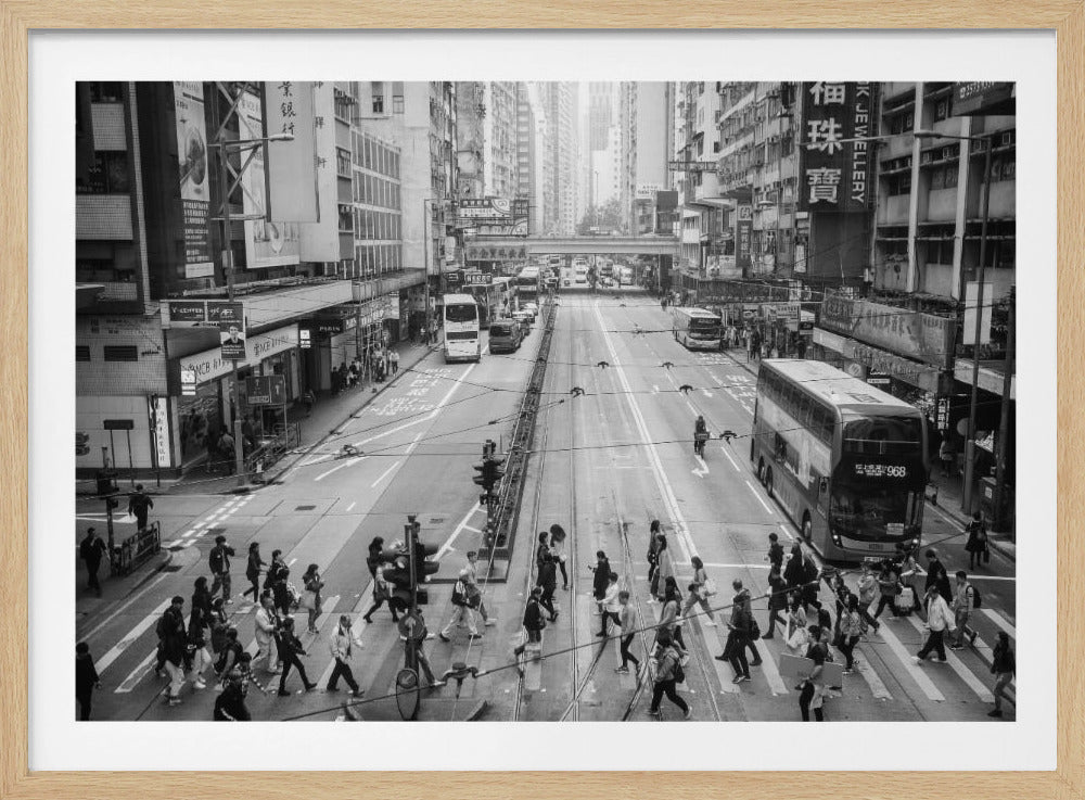 A framed, high-angle, black and white photograph of a bustling street scene in Hong Kong. The wide street is filled with vehicles, including double-decker buses, and numerous pedestrians are crossing at a crosswalk. The street is lined with tall buildings covered in signs with Chinese characters, creating a dense, energetic urban atmosphere. Print