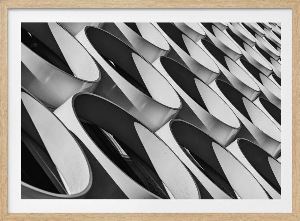 A framed, black and white photograph captures a low-angle, close-up view of a modern building's facade. The image features a repeating pattern of slanted, oval, or teardrop-shaped metallic elements, creating a dynamic interplay of light, shadow, and reflection. Print