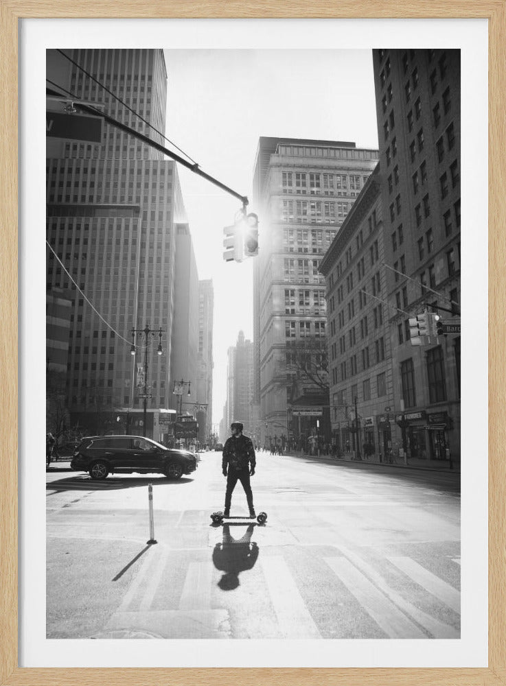 A black and white vertical photograph of a person on an electric skateboard, paused in the middle of a sun-drenched New York City street, with skyscrapers towering in the background and a long shadow cast forward. Print