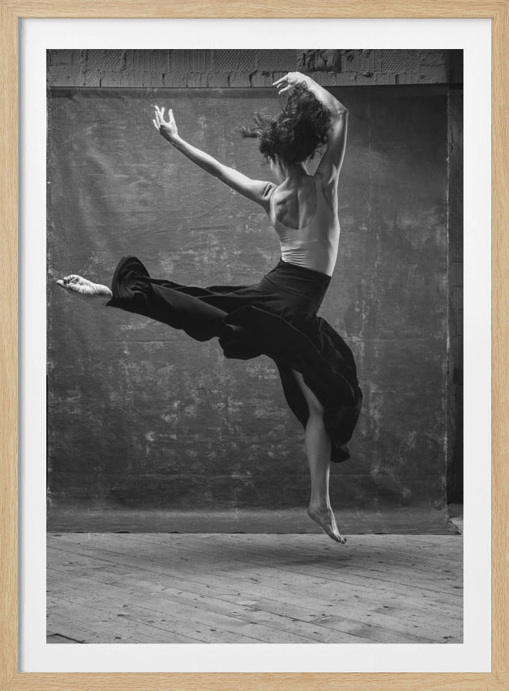 A dynamic black and white photograph of a female dancer captured mid-leap. Her back is toward the camera, showcasing her muscular form. She wears a light-colored tank top and a long, flowing black skirt that billows around her legs. Her arms are raised in an expressive pose, and she is suspended above a wooden floor against a mottled grey backdrop. Wall Art
