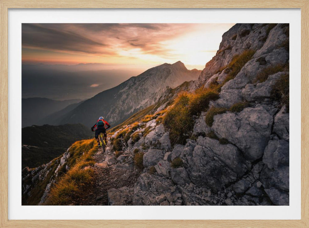 A mountain biker from behind rides along a narrow, rocky trail on a steep mountain ridge during a dramatic golden sunset. In the background, layers of mountains fade into the distance under a warm, glowing sky. Artwork