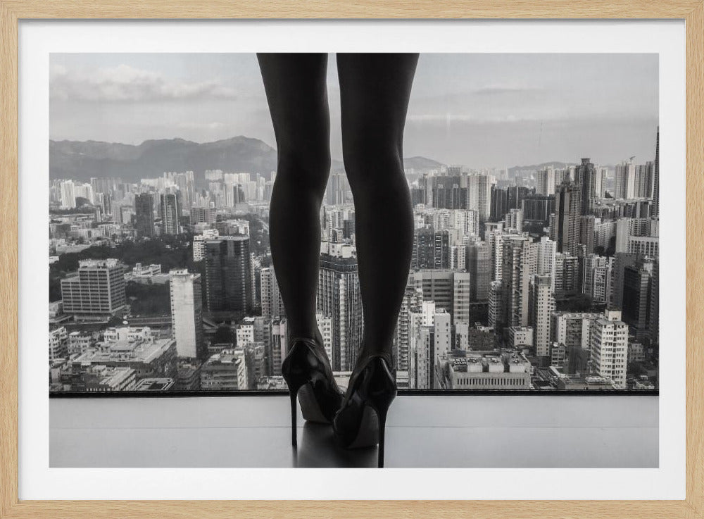 A framed black and white photograph showing the back of a woman's legs and high heels as she stands before a large window, looking out over a vast, dense cityscape with distant mountains under a cloudy sky. Artwork