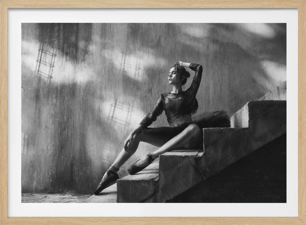 A dramatic black and white photograph of a graceful ballerina sitting on concrete stairs. She is wearing a dark lace top and pointe shoes, with her eyes closed and one hand behind her head in a serene pose, while shadows from a window play on the wall behind her. Wall Art