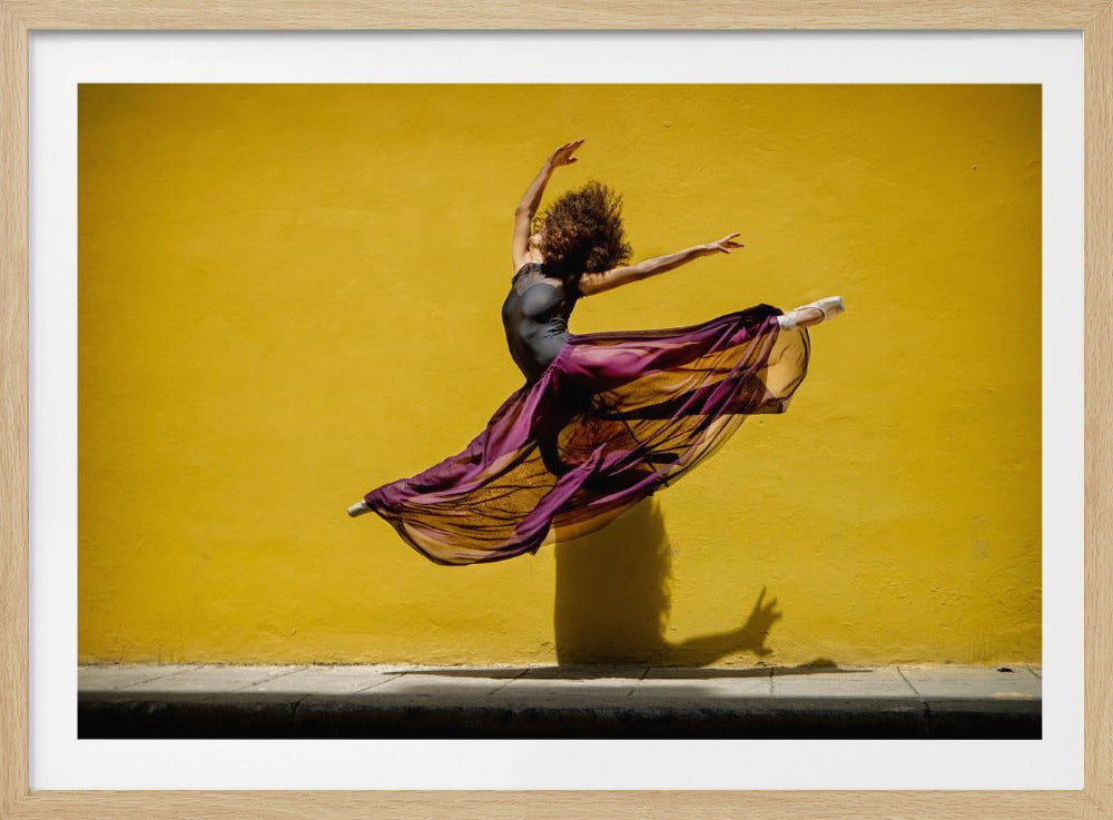 A framed photograph of a female ballet dancer with curly hair leaping gracefully in the air against a vibrant yellow wall. She wears a black leotard and a long, flowing sheer purple skirt that billows around her. A strong shadow is cast on the wall behind her. Poster