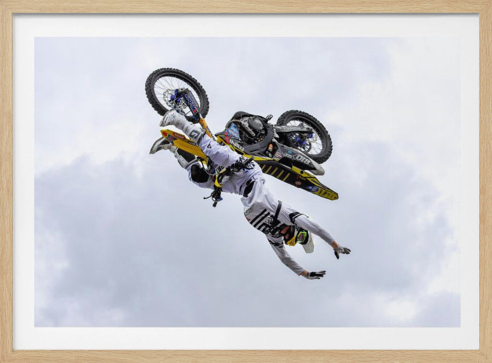 A framed action shot of a freestyle motocross rider in mid-air, performing an upside-down trick against a cloudy sky, with their body hanging below the yellow and black dirt bike. Artwork