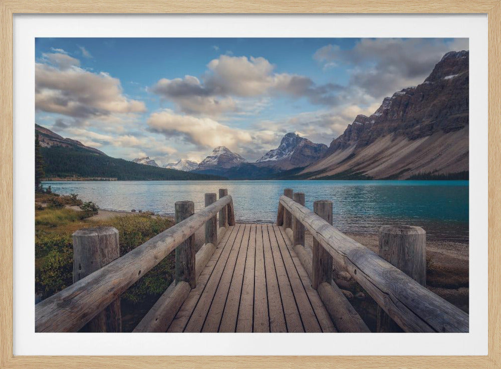 A wooden pier leads out onto a vibrant turquoise glacial lake, surrounded by towering, snow-dusted mountains under a dramatic cloudy sky. The image is presented in a silver frame. Artwork