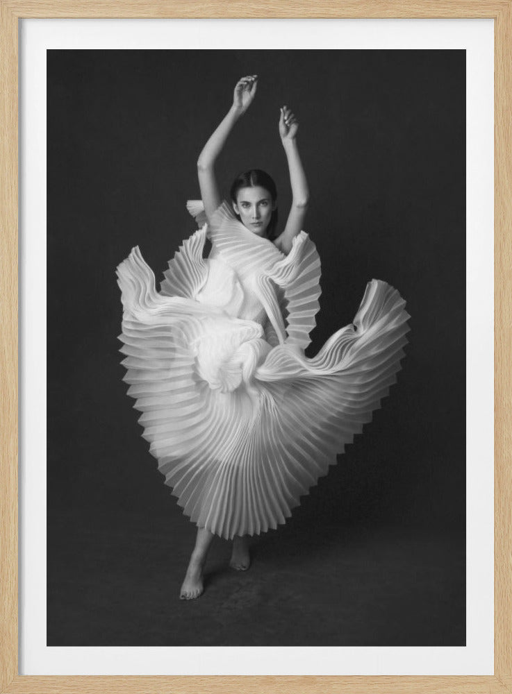 A framed, black and white photograph of a woman in a dramatic pose, wearing a voluminous white pleated dress. Her arms are raised above her head as the dress fans out around her in a sculptural, swirling motion. She looks directly at the camera against a solid dark gray background. Decor
