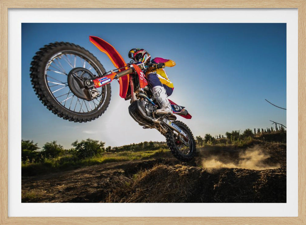 A dynamic, low-angle shot of a motocross rider in mid-air, jumping over a dirt mound. The rider is on a red, white, and blue dirt bike, wearing colorful racing gear and a helmet. The bike's front wheel is high in the air against a clear blue sky, while the back wheel is just clearing the jump, kicking up a cloud of dust. The background features a green, rural landscape. Wall Art