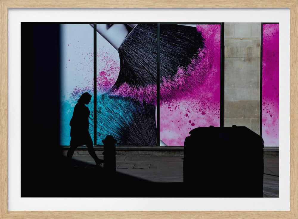 A framed photograph of a street scene where a person's silhouette is captured walking past a large, vibrant window advertisement. The ad features a close-up of a makeup brush with explosions of magenta and cyan powder. The foreground is cast in deep shadow, creating a dramatic contrast. Print