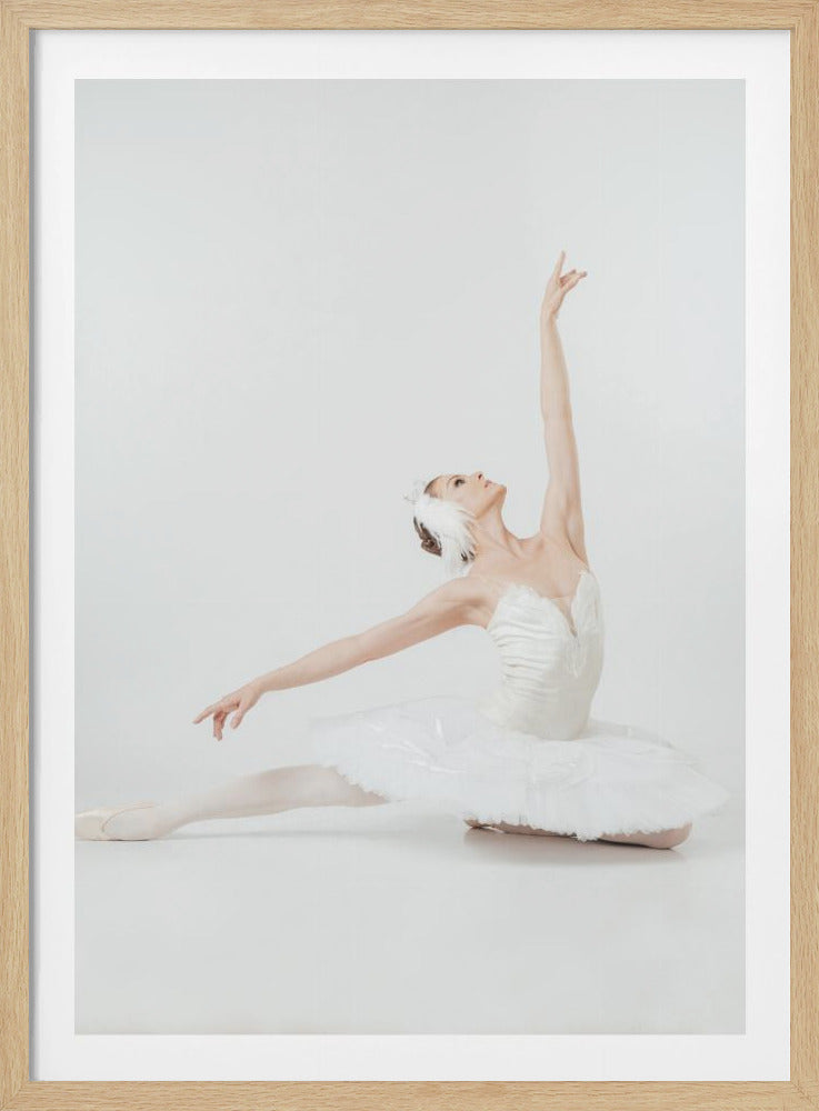 A framed photograph of a graceful ballerina dressed in a white swan costume, including a tutu and a feathered headpiece. She is posed on the floor against a plain white background, with one leg extended, her body arched back, and one arm reaching elegantly upwards. Wall Art