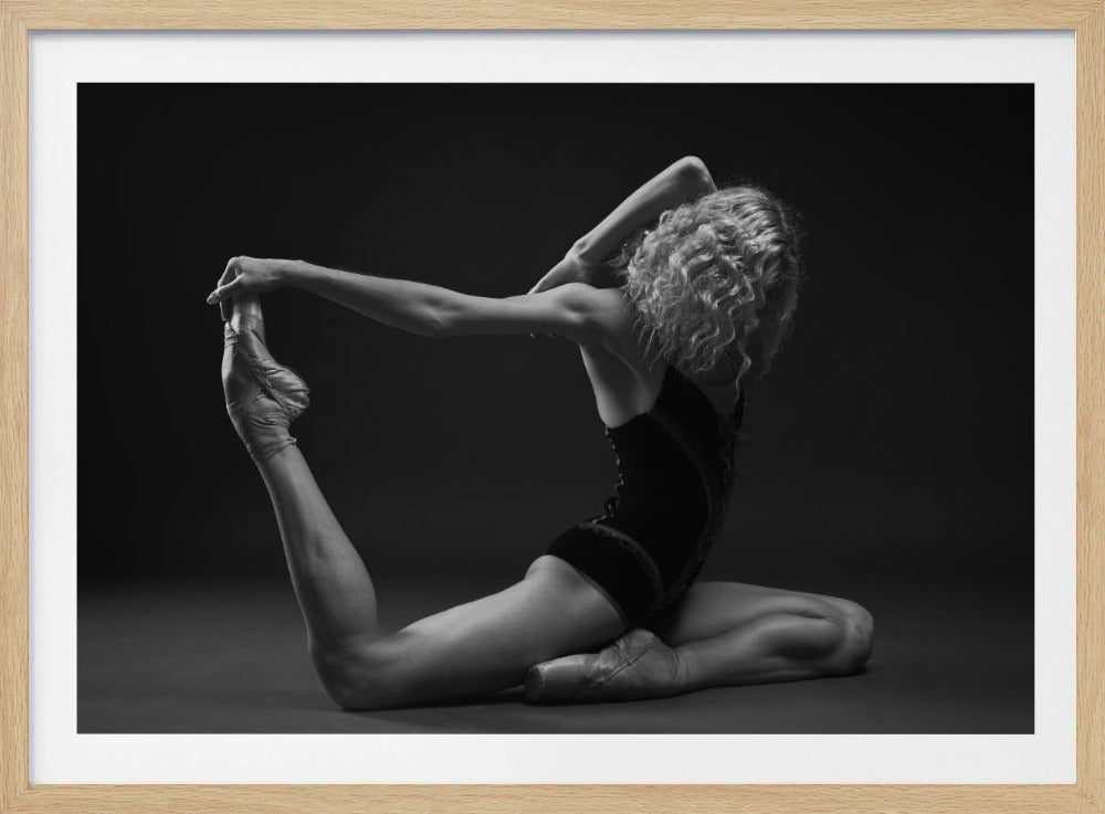 A black and white studio photograph of a flexible ballerina in a dark leotard and pointe shoes, arching her back to hold her extended leg over her head in an elegant pose against a dark background. Artwork