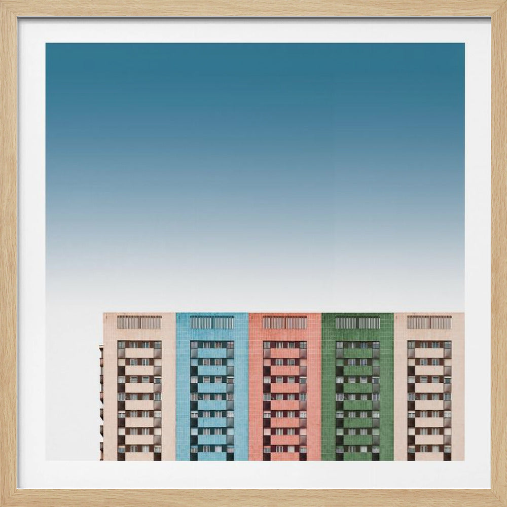 A framed minimalist photograph of a row of colorful apartment buildings set against a vast, clear blue sky. The buildings are identical in structure but colored beige, blue, red, and green, creating a vibrant, repeating pattern at the bottom of the frame. Decor