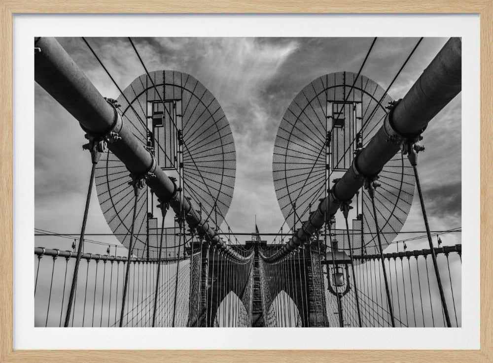 A symmetrical, low-angle black and white photograph of the Brooklyn Bridge's suspension cables and towers against a cloudy sky, framed in silver. Poster