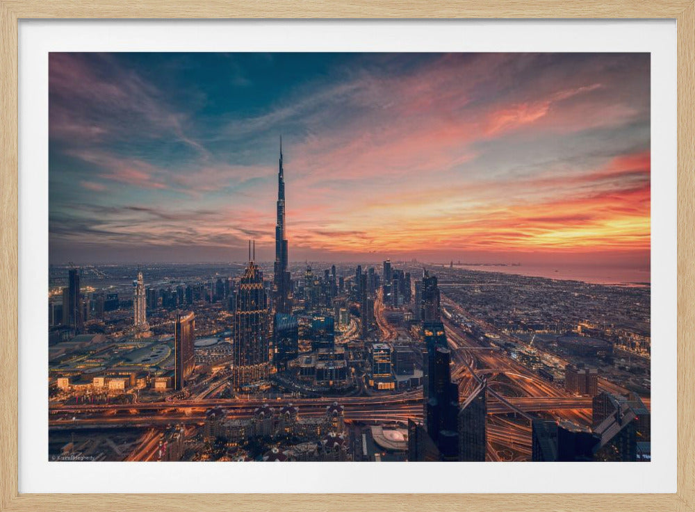 An aerial view of the Dubai skyline at sunset, with the Burj Khalifa prominently featured against a dramatic sky filled with orange, pink, and blue clouds. The sprawling city below is illuminated with golden lights from buildings and busy highways. Decor