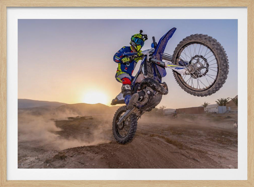 A motocross rider in blue and yellow gear is captured mid-air on a blue dirt bike, performing a jump against a dusty landscape with a setting sun in the background. The image is enclosed in a silver frame. Wall Art