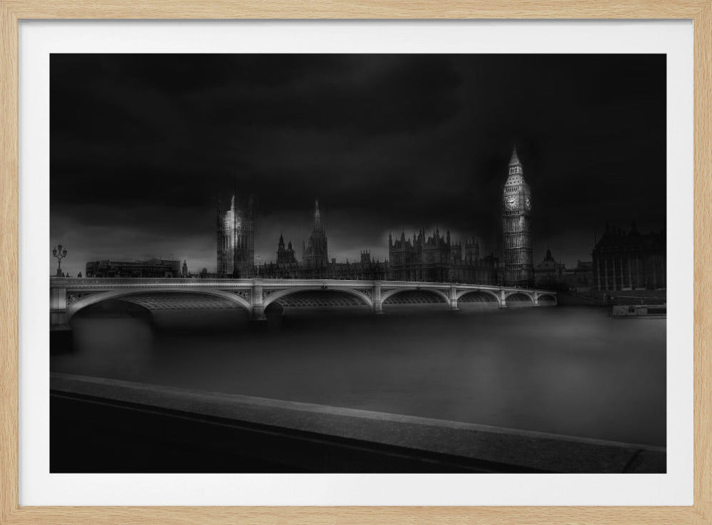 A moody, atmospheric black and white photograph of the Houses of Parliament and Big Ben in London, viewed from across the River Thames. The iconic buildings are softly illuminated against a dark, dramatic sky, with Westminster Bridge arching over the dark, smooth water in the foreground. The image is presented within a silver-colored frame. Artwork