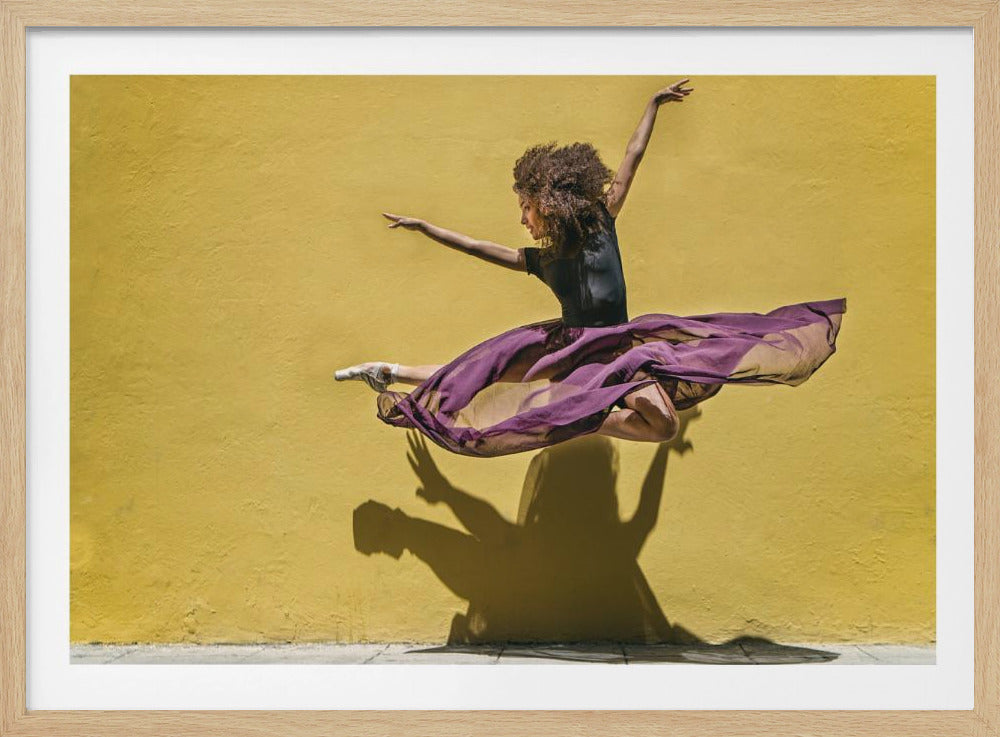 A framed photograph of a graceful ballerina with curly brown hair leaping in mid-air. She is wearing a black leotard and a flowing purple skirt against a vibrant, textured yellow wall, which casts a dramatic shadow of her pose. Wall Art