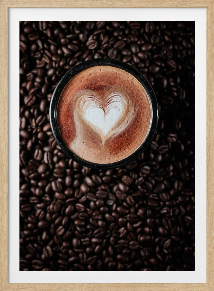 A top-down view of a cappuccino in a black mug with a white heart-shaped foam art design, placed on a bed of dark roasted coffee beans. Artwork