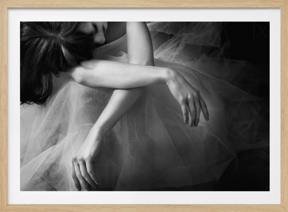 A black and white photograph of a woman with dark hair, seen from above, curled up on the floor. Her arms are crossed over her knees, and she is surrounded by the soft, translucent fabric of a tulle skirt. The mood is quiet and introspective. The photo is enclosed in a silver frame. Artwork