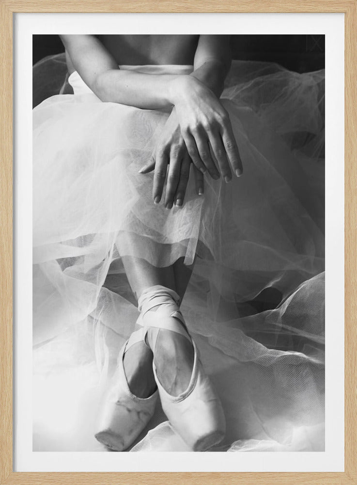 A black and white photograph of a seated ballerina, focusing on her hands resting on her white tulle tutu and her satin pointe shoes tied with ribbons. Wall Art