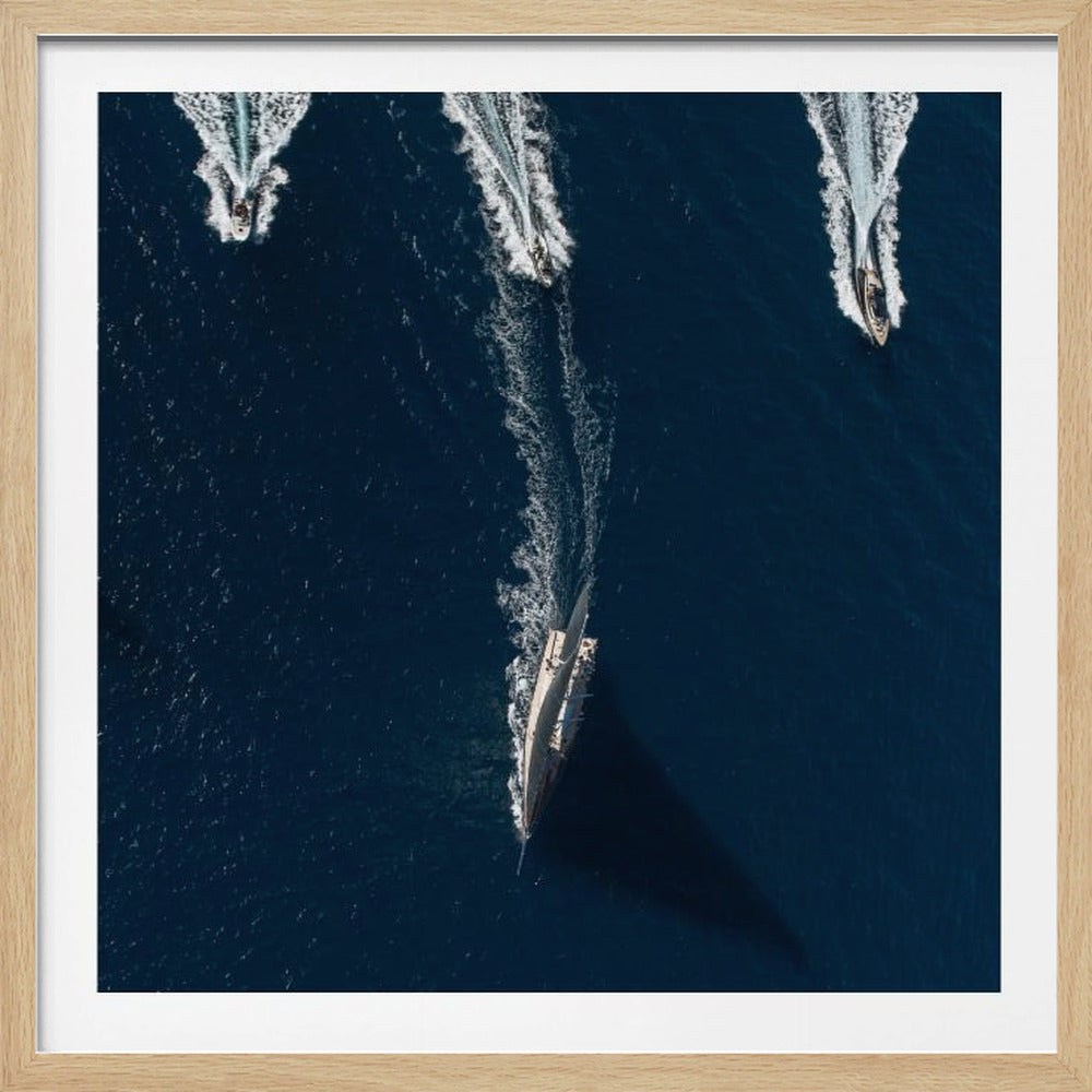 An aerial photograph from directly above shows a large sailboat and three smaller motorboats moving across a deep blue sea, leaving white wakes behind them. The sailboat is in the foreground, casting a long shadow, while the smaller boats are positioned further back. The entire image is enclosed in a light-colored wooden frame. Wall Art