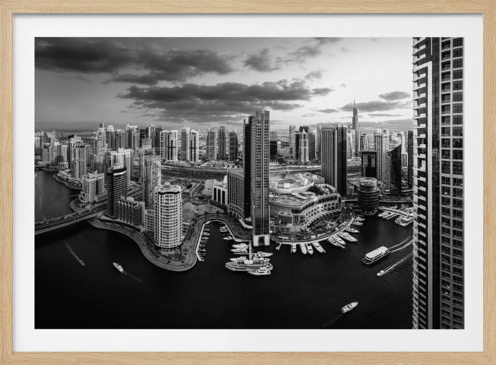 A framed, black and white panoramic photograph of the Dubai Marina taken from a high vantage point. The image captures the dense cityscape with numerous modern skyscrapers surrounding a winding waterway filled with docked yachts and moving boats. The sky is filled with dramatic, dark clouds, adding to the moody atmosphere of the urban landscape. Decor