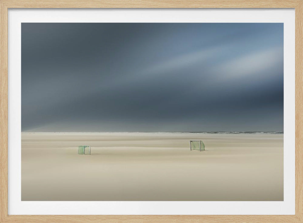 A minimalist long-exposure photograph of a vast, empty beach under a dark, moody sky. Two small, solitary soccer goals sit on the pale sand, with the ocean visible as a thin line in the distance. The artwork is presented in a silver frame. Artwork