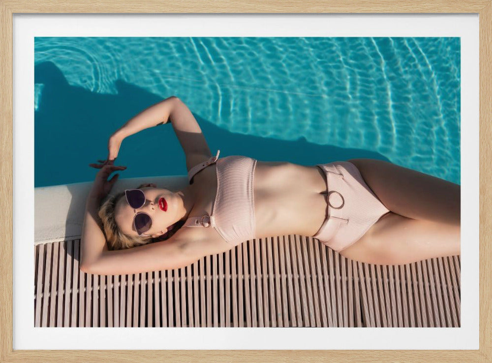An overhead shot of a stylish woman in a light pink bikini and sunglasses sunbathing on a slatted wooden deck beside the vibrant blue water of a swimming pool, all enclosed within a silver frame. Wall Art