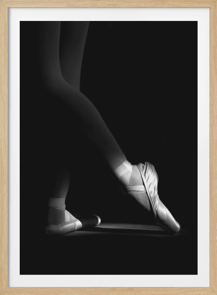 A dramatic black and white photograph showing the lower legs and feet of a ballet dancer, with one foot gracefully en pointe, highlighted by strong side lighting against a deep black background. Decor