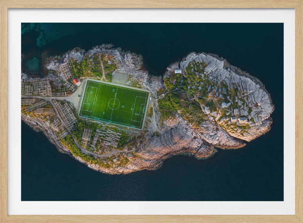 An aerial view of a vibrant green soccer field situated on a small, rocky island surrounded by dark blue water. The photograph, taken from directly above, captures the unique and remote location of the sports facility, with small buildings and wooden structures dotting the island's terrain. The entire scene is enclosed within a silver frame. Print