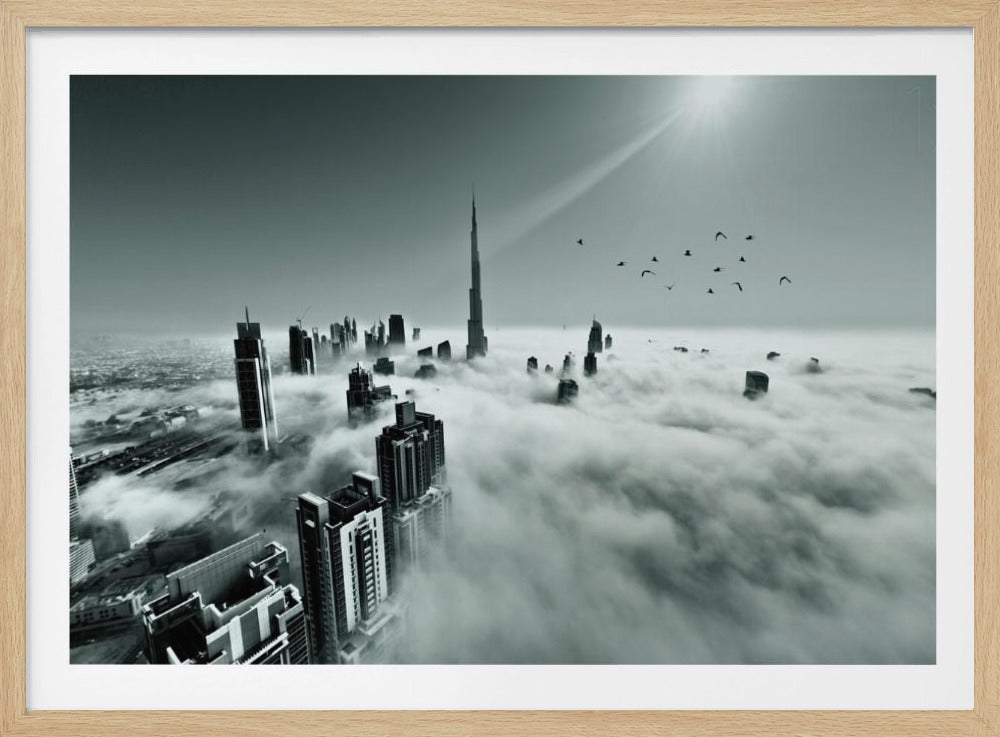 A framed, black and white aerial photograph of the Dubai skyline piercing through a thick layer of clouds. The Burj Khalifa stands prominently in the center, with other skyscrapers emerging from the fog. A flock of birds flies in the sky as a bright sunbeam shines down from the upper right corner. Wall Art